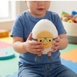 May include: A plush egg-shaped toy with a yellow yolk face, wearing blue glasses and smiling. The egg has blue feet and is held by a child wearing a blue striped shirt and jeans. The toy is soft and cuddly.