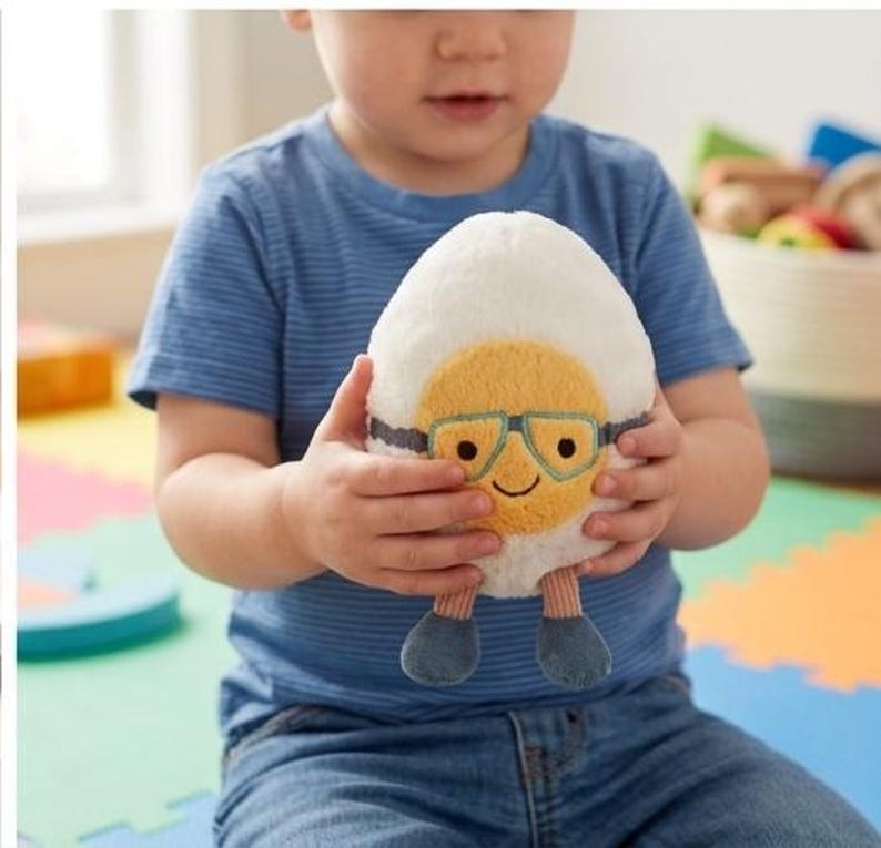 May include: A plush egg-shaped toy with a yellow yolk face, wearing blue glasses and smiling. The egg has blue feet and is held by a child wearing a blue striped shirt and jeans. The toy is soft and cuddly.