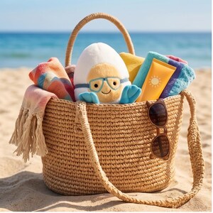 May include: A woven straw beach bag filled with beach essentials. Inside, a plush egg character wearing goggles sits among colorful towels, sunscreen, and sunglasses. The bag has a handle and a shoulder strap, perfect for a day at the beach.