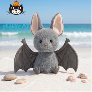 May include: A plush gray bat toy with large ears and outstretched wings, sitting on a sandy beach. The bat has a soft, furry texture and a cute, friendly expression. The background shows the ocean and sky.