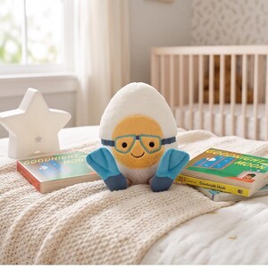 May include: A plush toy shaped like an egg with a yellow yolk face wearing blue goggles and flippers. The toy is sitting on a bed with two copies of the book "Goodnight Moon" and a star-shaped night light.
