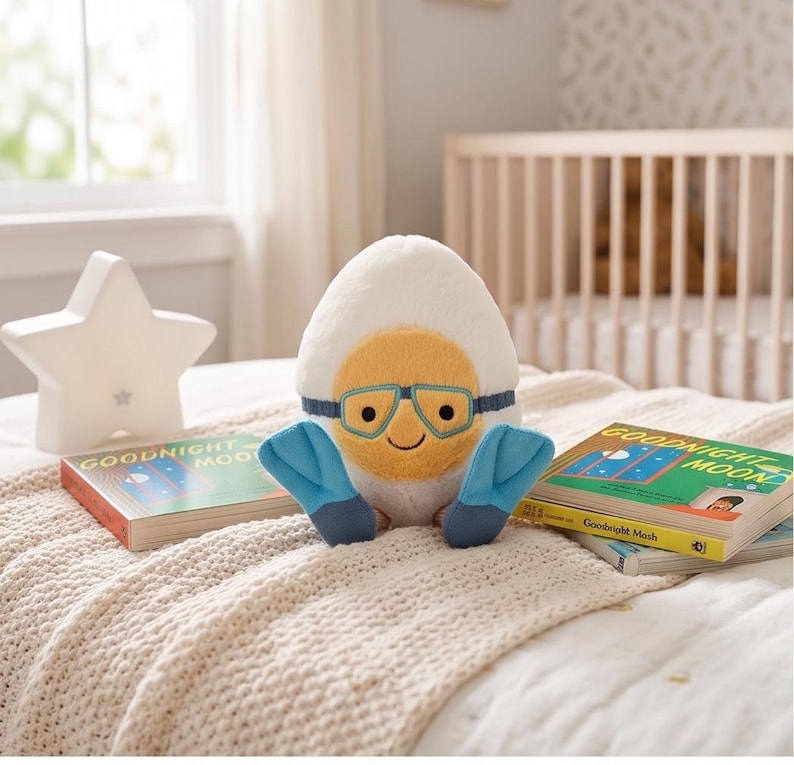 May include: A plush toy shaped like an egg with a yellow yolk face wearing blue goggles and flippers. The toy is sitting on a bed with two copies of the book "Goodnight Moon" and a star-shaped night light.