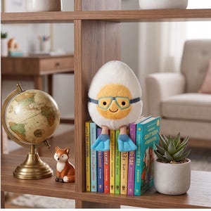May include: A plush egg character with glasses and flipper feet sits atop a stack of colorful books. A small globe and a fox figurine are on the shelf. The egg has a yellow yolk and a smiling face.