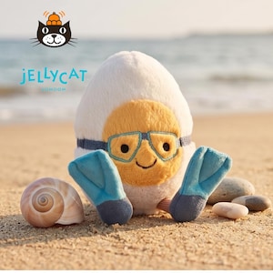 May include: A plush toy shaped like an egg, with a yellow face wearing blue goggles and a blue strap. It has blue flippers and is on a sandy beach. The brand name "Jellycat London" is visible.