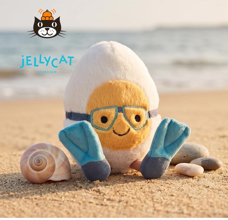 May include: A plush toy shaped like an egg, with a yellow face wearing blue goggles and a blue strap. It has blue flippers and is on a sandy beach. The brand name "Jellycat London" is visible.