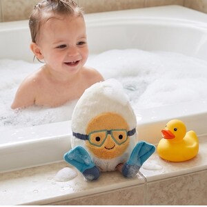 May include: A plush, white egg-shaped bath toy with a smiling face, blue goggles, and blue flipper feet sits next to a yellow rubber duck. A baby is in a bathtub filled with bubbles.