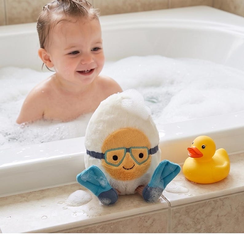 May include: A plush, white egg-shaped bath toy with a smiling face, blue goggles, and blue flipper feet sits next to a yellow rubber duck. A baby is in a bathtub filled with bubbles.
