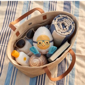 May include: A beige tote bag with brown leather handles, filled with beach essentials. Includes sunglasses, sunscreen, a rolled blue and white striped towel, a book, a seashell, and a plush egg character wearing goggles and flippers.