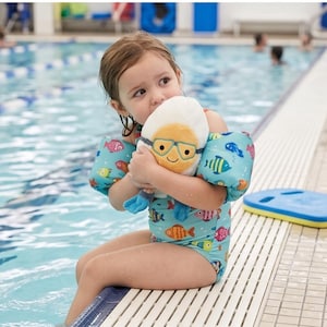 May include: A young child wearing a turquoise swimsuit and arm floaties, hugging a plush toy shaped like a swimmer. The swimsuit and floaties feature colorful fish illustrations. The child is sitting by a swimming pool.