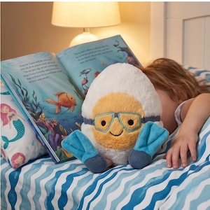 May include: A plush toy shaped like a diver's helmet with a smiling face, goggles, and flippers. The toy is white, yellow, and blue, resting on a bed with a mermaid pillow and an open book with illustrations of marine life.