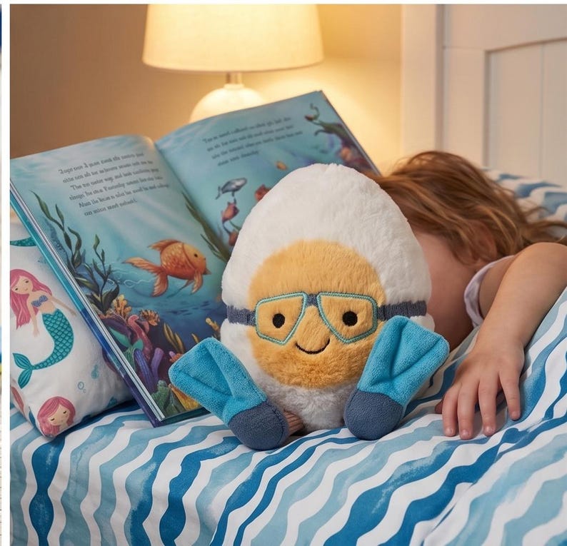 May include: A plush toy shaped like a diver's helmet with a smiling face, goggles, and flippers. The toy is white, yellow, and blue, resting on a bed with a mermaid pillow and an open book with illustrations of marine life.