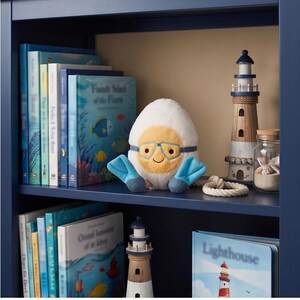 May include: A navy blue bookshelf displays children's books, a plush egg character with goggles and flippers, and decorative lighthouse figurines. A glass jar with seashells and a rope accent complete the nautical theme.
