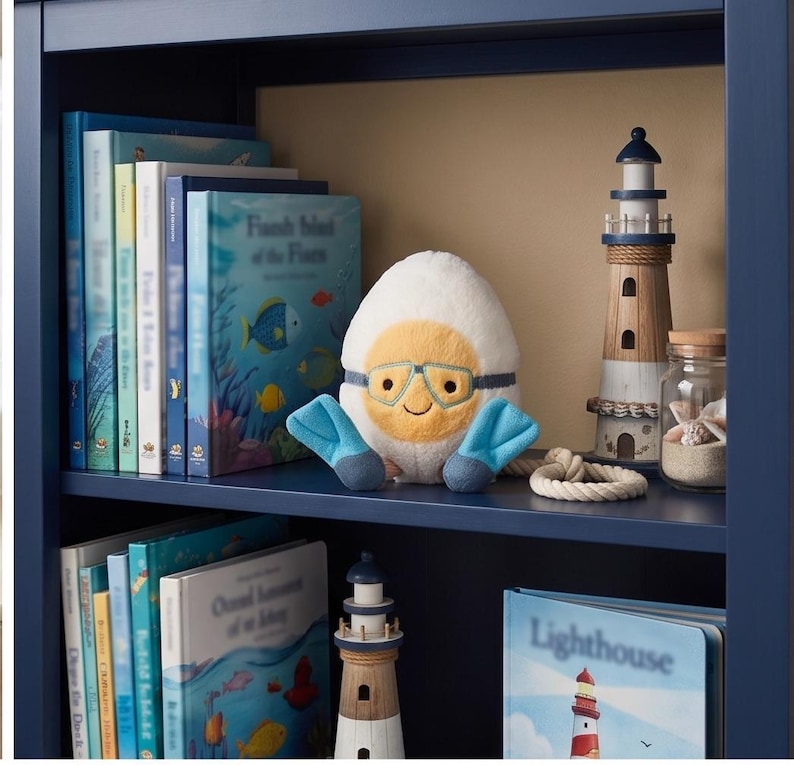 May include: A navy blue bookshelf displays children's books, a plush egg character with goggles and flippers, and decorative lighthouse figurines. A glass jar with seashells and a rope accent complete the nautical theme.