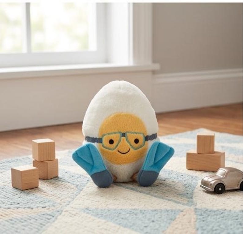 May include: A plush egg-shaped toy with a yellow face wearing teal-rimmed glasses and blue flipper-like appendages. The toy is on a patterned rug with wooden blocks and a toy car nearby. The egg has a smiling face.
