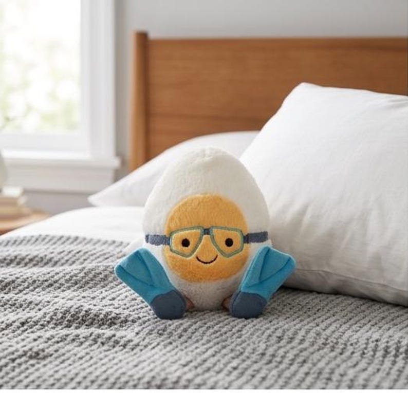 May include: A plush egg-shaped toy with a smiling yellow face, blue goggles, and blue flippers. The egg is white with a yellow yolk-like center. The toy is sitting on a gray blanket on a bed.