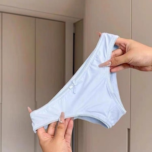 May include: Pale blue underwear with a small bow. The edges are scalloped. The garment is held up by two hands. The background features a light-coloured wall and a closed door.