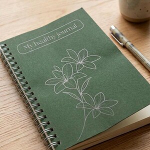Puede incluir: Un diario verde con espiral con el texto "My healthy journal" y un dise&ntilde;o floral blanco. El diario est&aacute; abierto, revelando p&aacute;ginas en blanco. Un bol&iacute;grafo y una taza est&aacute;n sobre la superficie de madera.