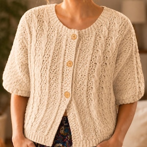 Peut inclure: Un gilet en tricot crème à manches courtes avec une encolure ronde, des motifs de torsades verticales et trois boutons en bois brun clair sur le devant. Le vêtement présente un motif texturé et des bords-côtes au col et aux poignets.