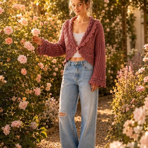 May include: A dusty rose crochet cardigan featuring a scalloped collar and wide sleeves, fastened with a single button at the center. The garment is paired with light blue denim jeans that have a distressed knee detail and a white tank top.