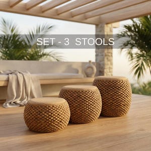 May include: Three round woven stools with tan cushions are arranged on a wooden deck. The stools have a textured, basket-weave design in a natural brown color. The text "SET - 3 STOOLS" is displayed above the stools.