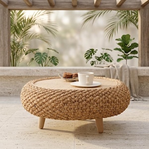 May include: A round, woven wicker coffee table with a light wood top and legs. A white teacup and saucer sit on the table, along with a small plate of cookies. The table is outdoors, with greenery in the background.