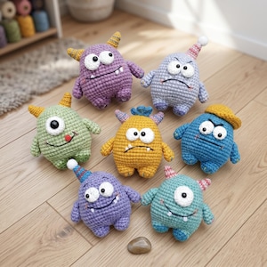 Puede incluir: Una colección de siete muñecos de monstruos de ganchillo de colores. Cada muñeco tiene una expresión única, cuernos y algunos llevan gorros de fiesta. Los muñecos son de tonos morado, azul, verde y amarillo, y están colocados sobre un suelo de madera.