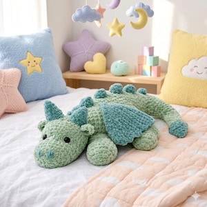 May include: A handmade, green crochet dragon plush toy with teal accents, lying on a bed. The dragon has horns, wings, and a long tail. Soft pillows and a mobile with stars, clouds, and moons are in the background.