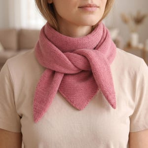 May include: A knitted scarf in a dusty rose hue is wrapped around the neck. The scarf is made from soft, textured yarn and knotted at the front. The ends of the scarf form a triangle. The background is blurred.