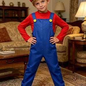 Pu&ograve; includere: Un bambino indossa un cappello rosso con una "M" bianca, una maglietta rossa a maniche lunghe e una tuta blu con bottoni gialli. Il bambino indossa anche stivali marroni. L'outfit assomiglia a un popolare personaggio dei videogiochi.