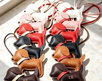 Puede incluir: Una colección de llaveros de cuero con forma de perro en varios colores, incluyendo blanco, rosa, rojo, negro, bronceado y marrón oscuro. Cada llavero cuenta con un collar en contraste y una correa fina. Dos llaveros oscuros tienen grabadas las letras RE y LK, mientras que otro tiene grabadas las letras KU.