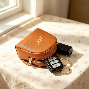May include: A small, tan, semi-circular zipped pouch with a textured surface and the gold-embossed letters BK on the front. The pouch is partially open, revealing a black car key fob and a gold-toned metal key ring inside the main compartment.