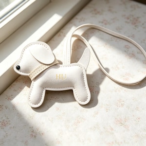 Puede incluir: Un bolso con forma de perro de color crema con una correa larga, que presenta una peque&ntilde;a cuenta negra como ojo, un detalle de collar y las letras HU grabadas en dorado en el lateral. El accesorio est&aacute; construido con costuras visibles en los bordes.