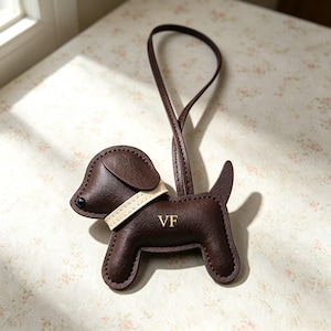 Puede incluir: Un colgante para bolso con forma de perro en color marr&oacute;n oscuro, con un collar color crema, un peque&ntilde;o ojo de cuenta negra y una correa larga. El cuerpo del perro tiene grabadas las letras doradas VF y presenta costuras visibles.