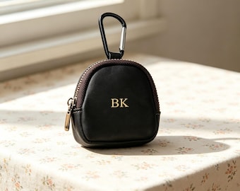 Puede incluir: Un pequeño estuche negro en forma de cúpula con una cremallera marrón oscuro y un mosquetón negro sujeto en la parte superior. La parte frontal del estuche presenta las letras doradas BK grabadas en el centro. Tiene un acabado mate liso.