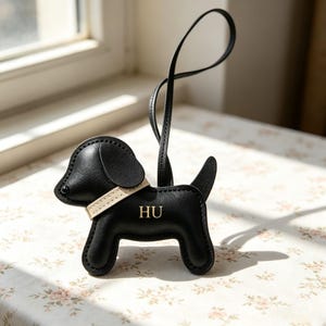 May include: A black leather dog-shaped bag charm featuring a light beige collar and the letters HU embossed in gold on its side. The charm includes a long, thin black leather strap for attachment, with visible stitching along the edges of the dog.