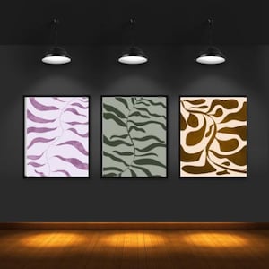 Peut inclure: Trois impressions d'art abstrait encadrées avec des cadres noirs sont accrochées à un mur gris foncé. Les impressions présentent des motifs organiques et fluides dans des tons de violet, de vert et de marron. La pièce est éclairée par trois spots et possède un sol en bois.
