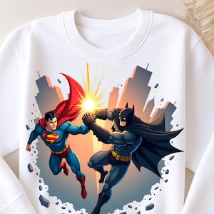 May include: A white crewneck sweatshirt featuring a graphic illustration of Superman and Batman in mid-air, touching fists to create a bright yellow light burst. The background depicts a stylized city skyline with floating debris and rocks.