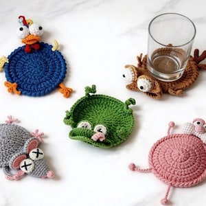 May include: A collection of five handmade crochet coasters in whimsical animal designs. Includes a blue chicken, brown crab, green frog, gray mouse, and pink snail. Each coaster features detailed facial features and is designed to hold a glass or mug.