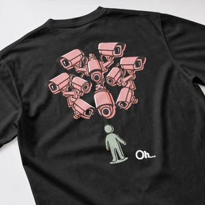 Puede incluir: Una camiseta negra con un estampado gráfico en la espalda. El diseño muestra un grupo de cámaras de seguridad rosas apuntando hacia abajo a una pequeña figura de palo de color verde claro. El texto "Oh..." está impreso en blanco al lado.