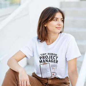 Può includere: Una t-shirt bianca a maniche corte con il testo nero TINY HUMAN PROJECT MANAGER sopra la grafica di una cartellina con una lista di cose da fare e una tazza di caffè con un cuore. Il testo REQUESTS ACCEPTED 24/7 è stampato accanto alla tazza.