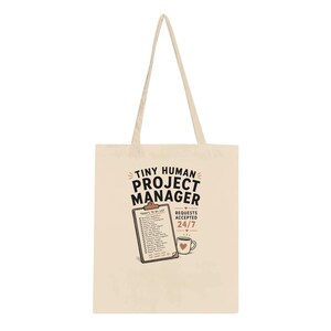 Può includere: Una borsa tote in tela beige con una stampa grafica che riporta il testo "TINY HUMAN PROJECT MANAGER" sopra una cartellina con una lista di cose da fare e una tazza di caffè con un'icona a forma di cuore. Il testo "REQUESTS ACCEPTED 24/7" è stampato accanto.