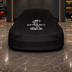 以下が含まれることがあります： 白い「Shelby」のロゴとコブラのエンブレムが付いた黒い車のカバー。 ショールームの車にかぶせられており、背景には他の車が見えます。 床は磨かれた濃い灰色の大理石です。