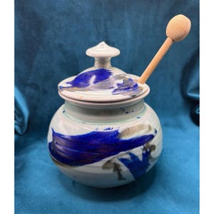 May include: A ceramic honey pot with a lid and a wooden honey dipper. The pot is white with blue and gray abstract brushstrokes. The lid is white with a blue and gray design. The honey dipper is made of light-colored wood.