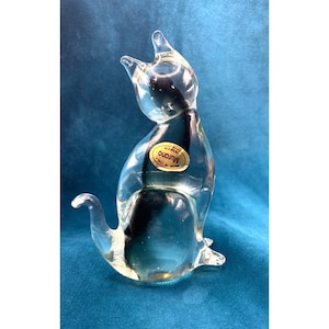 Op de afbeelding: Een helder glazen kattenbeeldje in zittende houding met de staart opzij gekruld. Het figuurtje heeft een gouden ovale sticker op de borst met de tekst Murano Made in Italy. Het glas is transparant en glad met duidelijk gevormde oren.