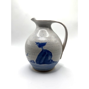 May include: A ceramic pitcher with a speckled, light gray finish. A blue bird is painted on a blue rock. The pitcher has a curved handle and a spout for pouring. The base of the pitcher is light blue.