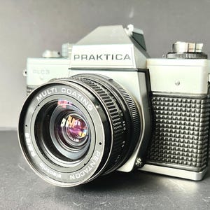 May include: A silver Praktica PLC3 35mm film camera featuring a black Pentacon electric 2.8/29 multi-coating lens. The camera body has a textured black grip on the side, a silver top plate with dials, and the brand name printed in black letters.