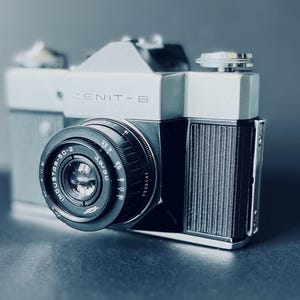 May include: A silver and black Zenit-E 35mm film camera featuring an Industar-50-2 3.5/50 lens. The camera body has a textured black grip on the right side, a silver top plate with the Zenit-E logo, and a prominent circular lens barrel with white text.
