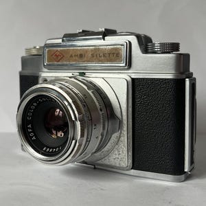 May include: An Agfa Ambi Silette 35mm film camera with a silver metal body and black textured leatherette grip. The front features a Color-Solinar lens with visible aperture and focus rings, and a rectangular nameplate reading AMBI SILETTE.