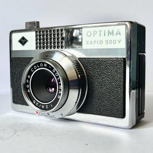 May include: An Agfa Optima Rapid 500 V camera with a chrome and black textured body. The front features a lens labelled Agfa Color-Solinar 1:2.8/35, a viewfinder, and a flash sync port. The top right corner displays the text Optima Rapid 500 V.