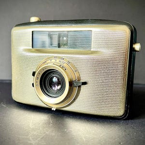 May include: A vintage film camera with a textured gold-tone front panel and a black body. The lens barrel is marked with the text DOMIPLAN and features adjustable settings for aperture and shutter speed, including numbers 30, 60, 125, 8, 11, 16, and 22.